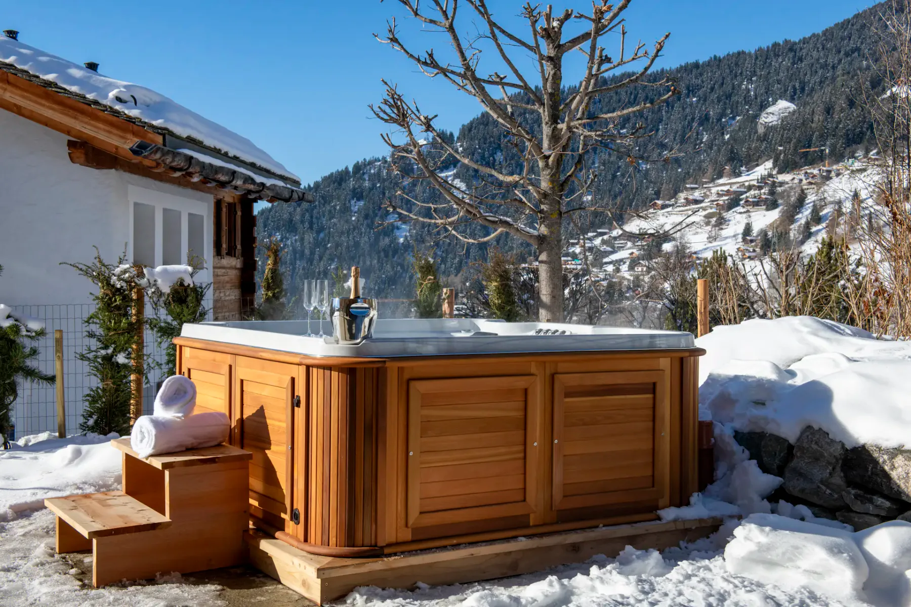 A snowy mountain landscape with a hot tub in the foreground, wooden chalets, and a clear blue sky with the sun shining brightly.