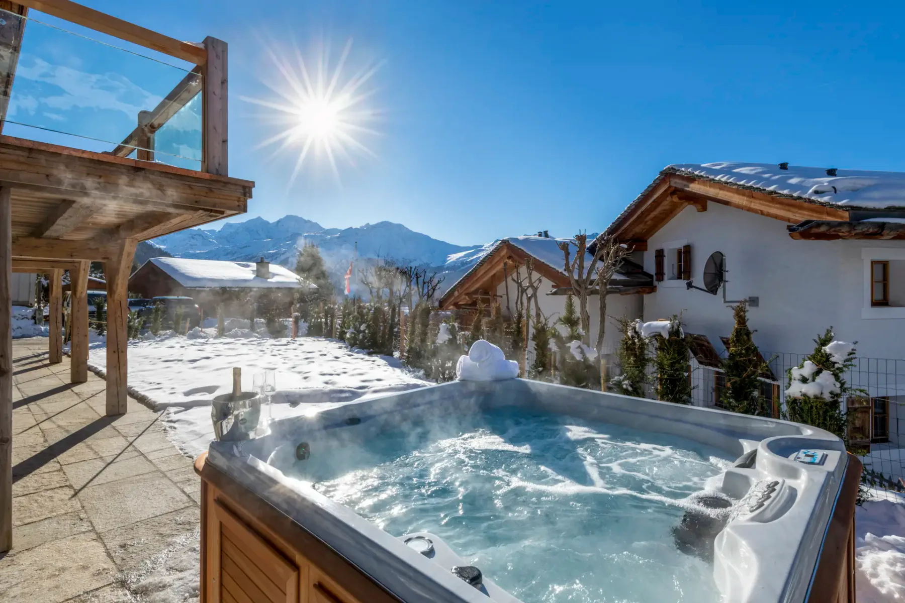 A snowy mountain landscape with a hot tub in the foreground, wooden chalets, and a clear blue sky with the sun shining brightly.