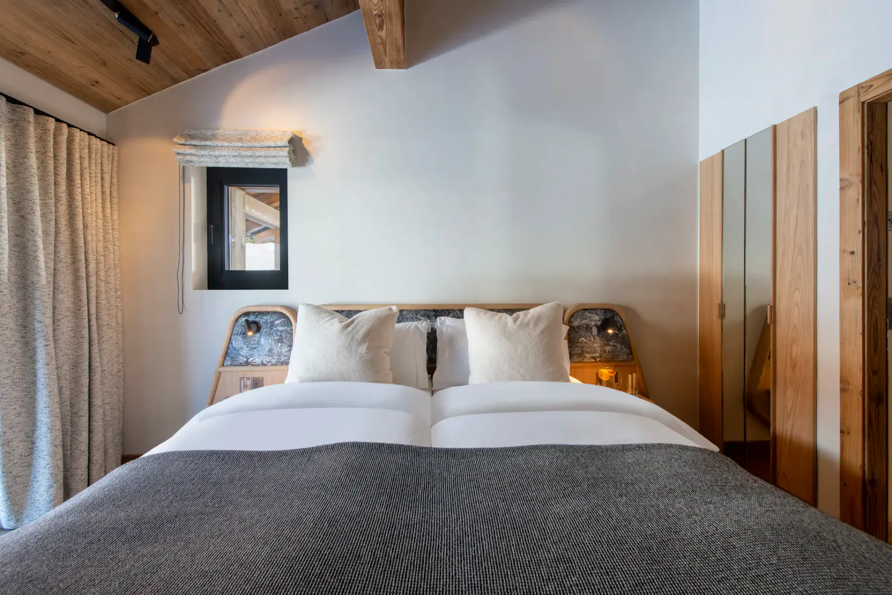 Modern bedroom with wooden ceiling and floor, featuring a double bed with white linens, gray throw, and two pillows. There's a large window with curtains and a wall-mounted flat-screen TV.