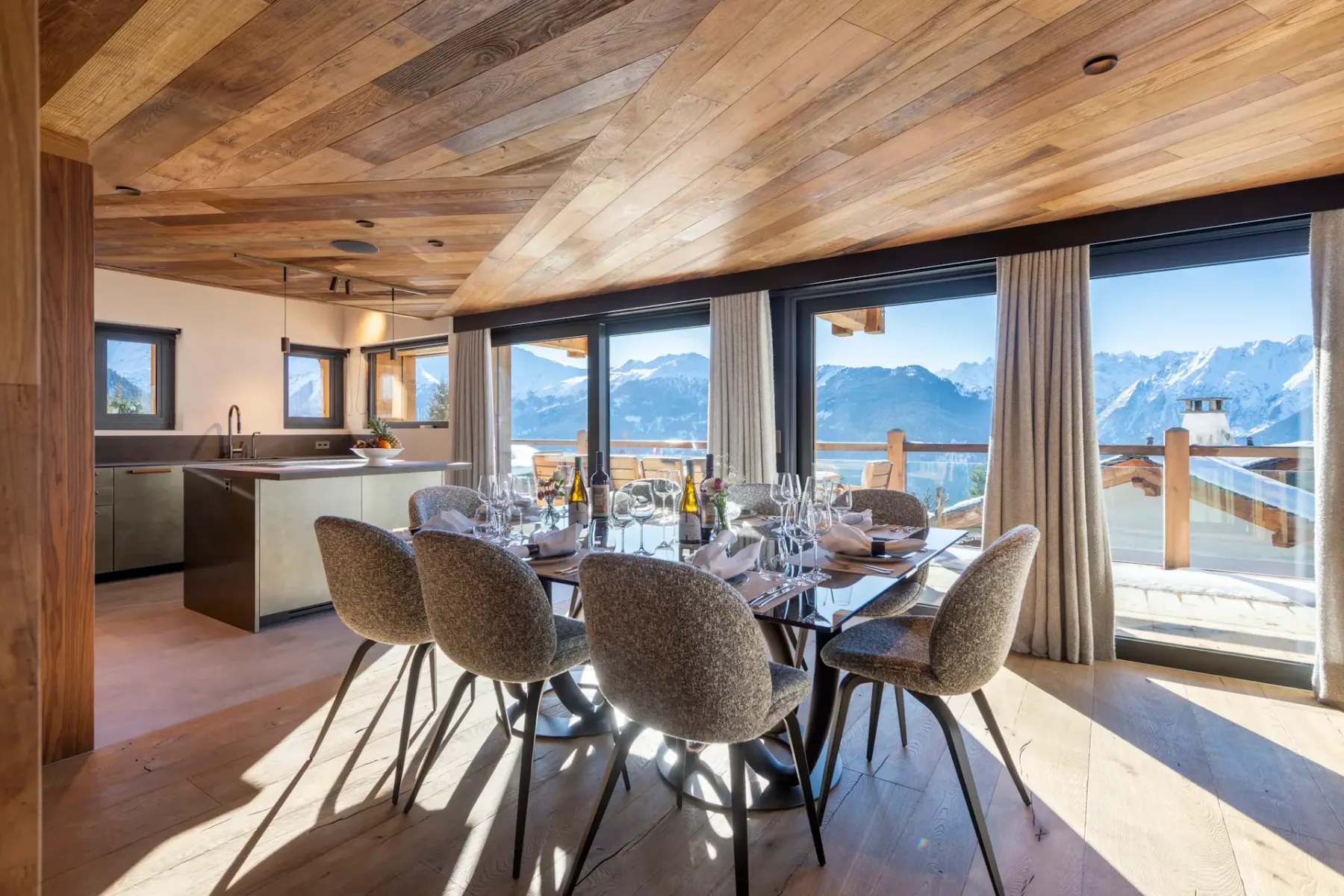 Modern dining area with a wooden ceiling, large windows showcasing a mountain view. A set table with chairs is in the foreground and a kitchen is visible in the background.