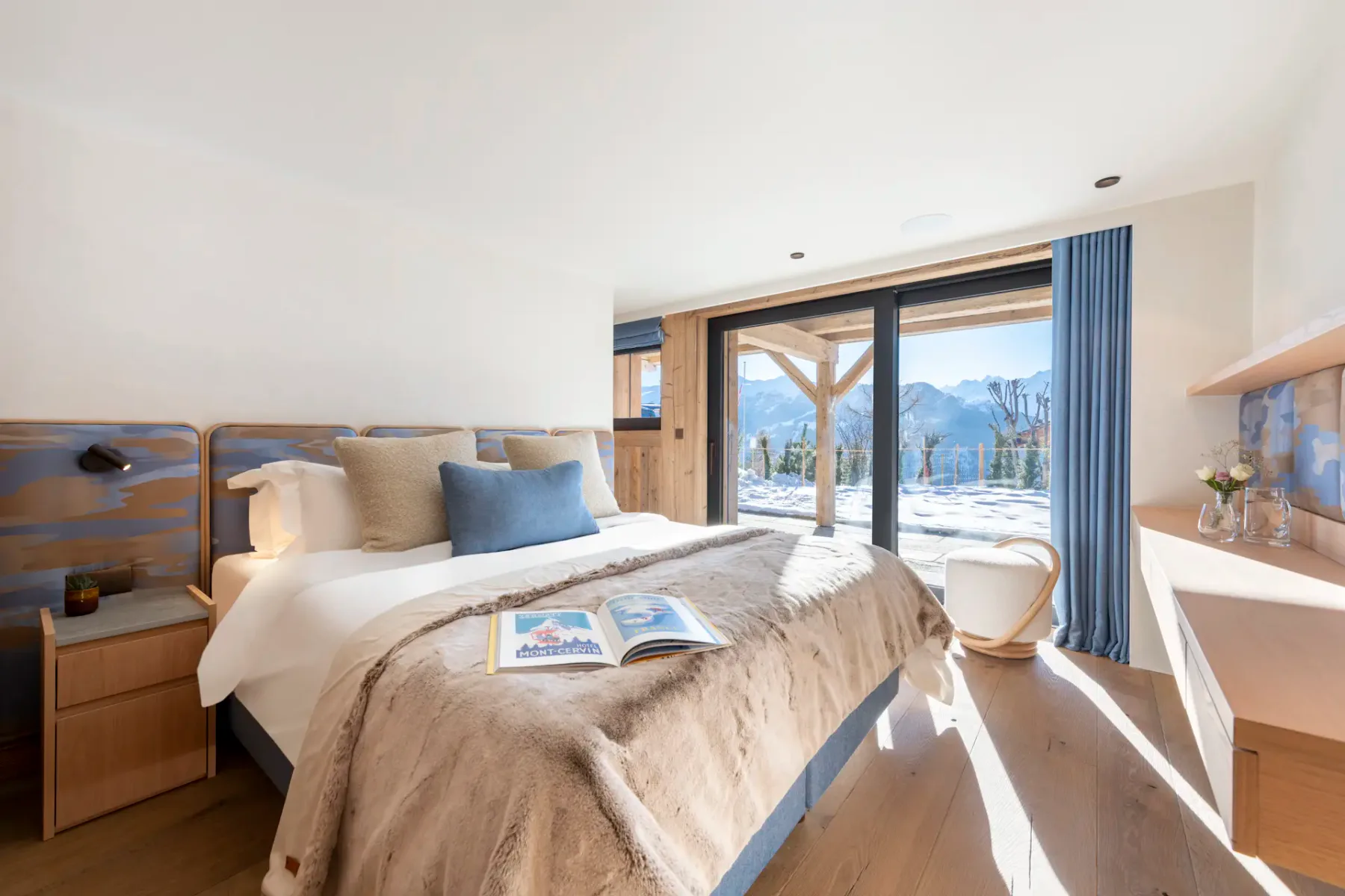 A bright bedroom with a double bed, beige and blue decor, bedside tables, and a scenic mountain view through large sliding glass doors. An open magazine rests on the bed.