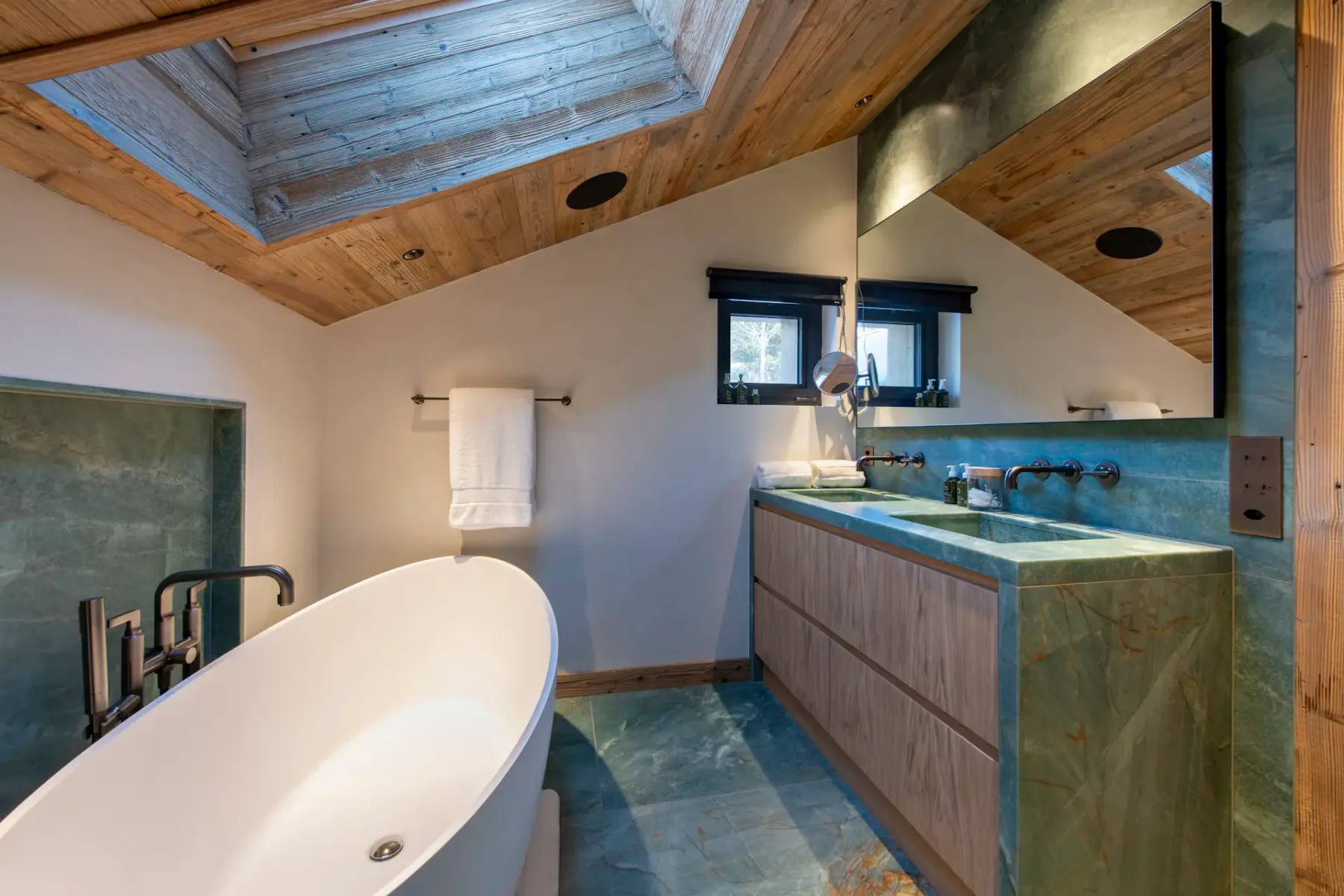 Modern bathroom with a freestanding white bathtub, wooden ceiling, skylight, dual-sink vanity with mirrors, and green stone countertops.