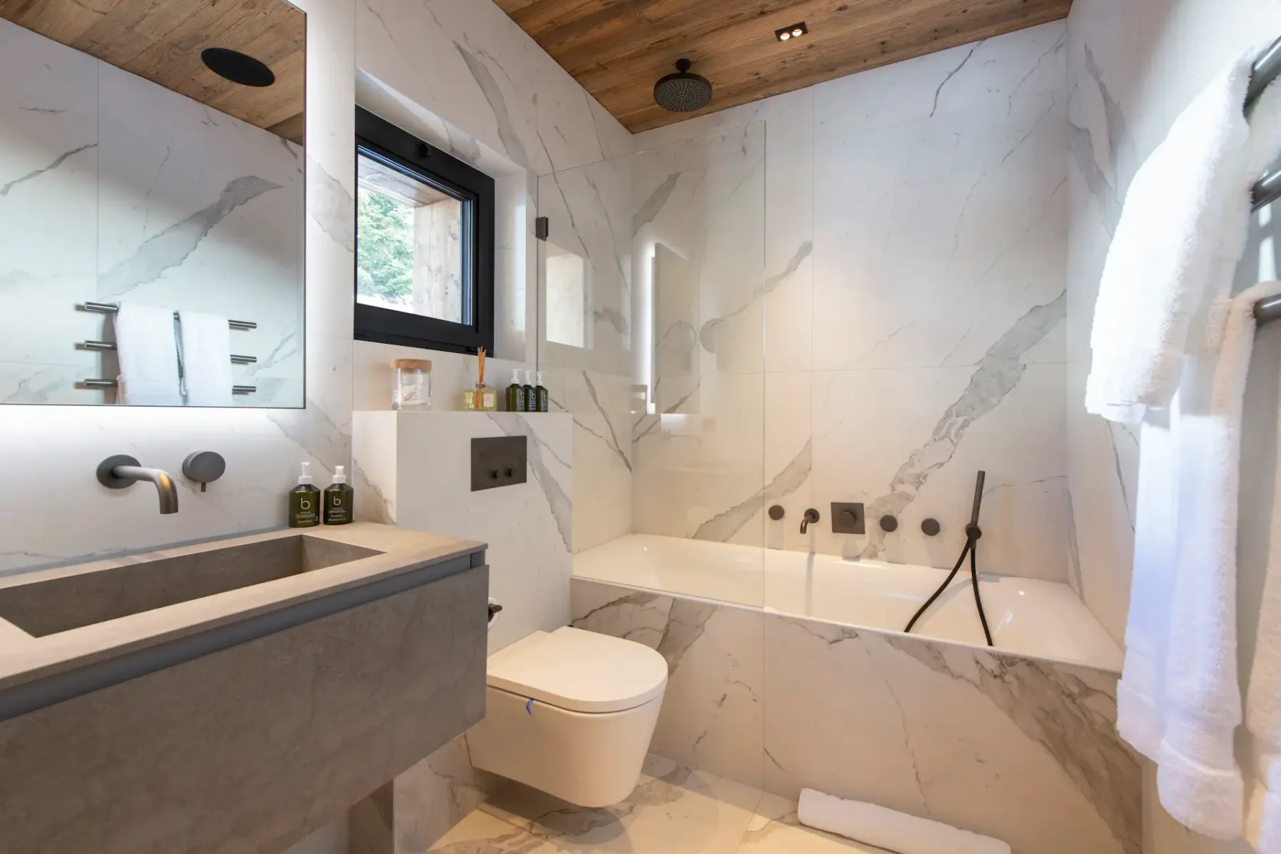 Modern bathroom with marble walls, a bathtub, overhead shower, wall-mounted sink, and toilet. A window is above the bathtub, and towels hang on a rail beside the sink.