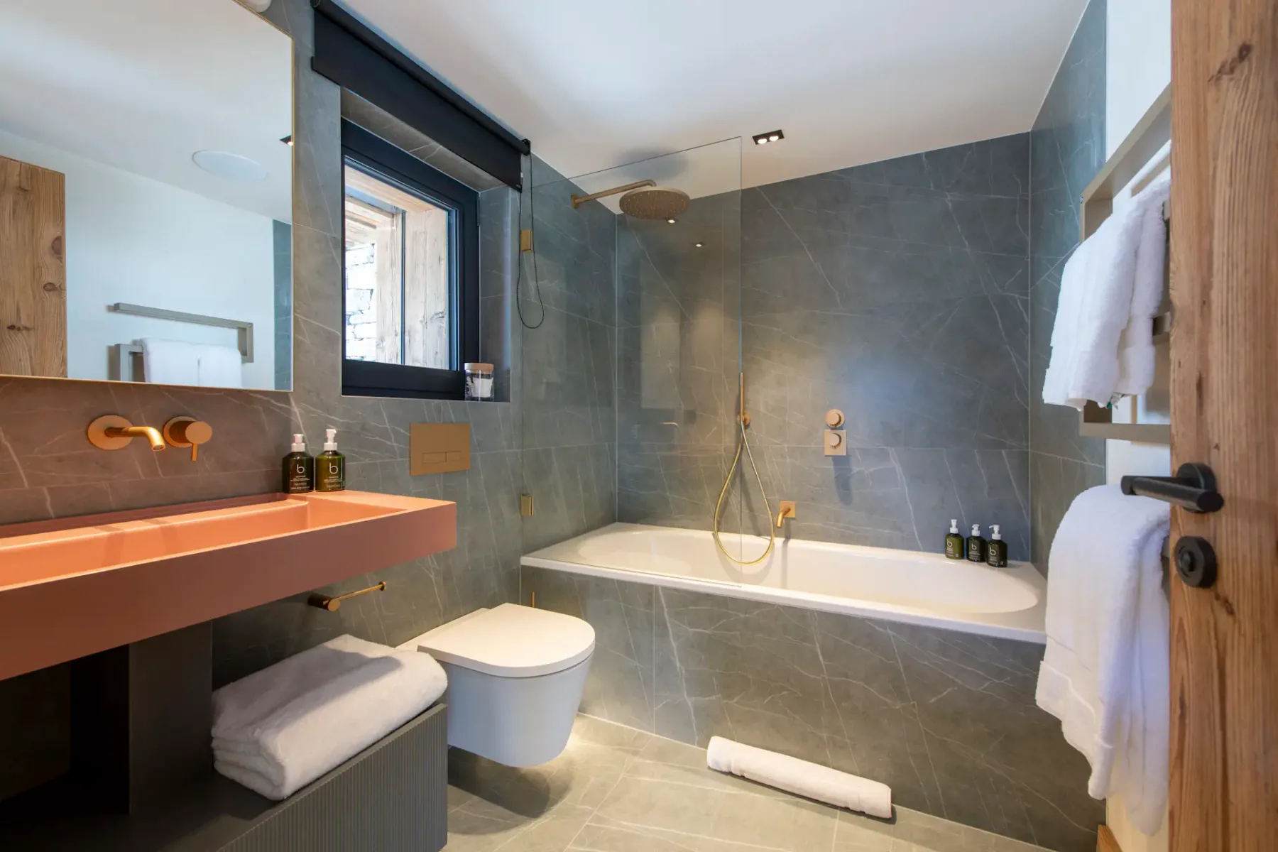 Modern bathroom with gray tile walls, a bathtub with a glass shower partition, wall-mounted toilet, and a pink countertop sink. Towels and toiletries are neatly arranged.