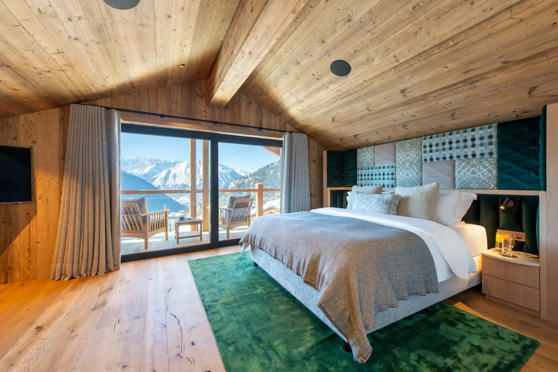 A cozy bedroom with wooden ceiling and floors, a large bed, and a balcony offering a mountain view. Two chairs are on the balcony, and light-colored curtains frame the sliding doors.