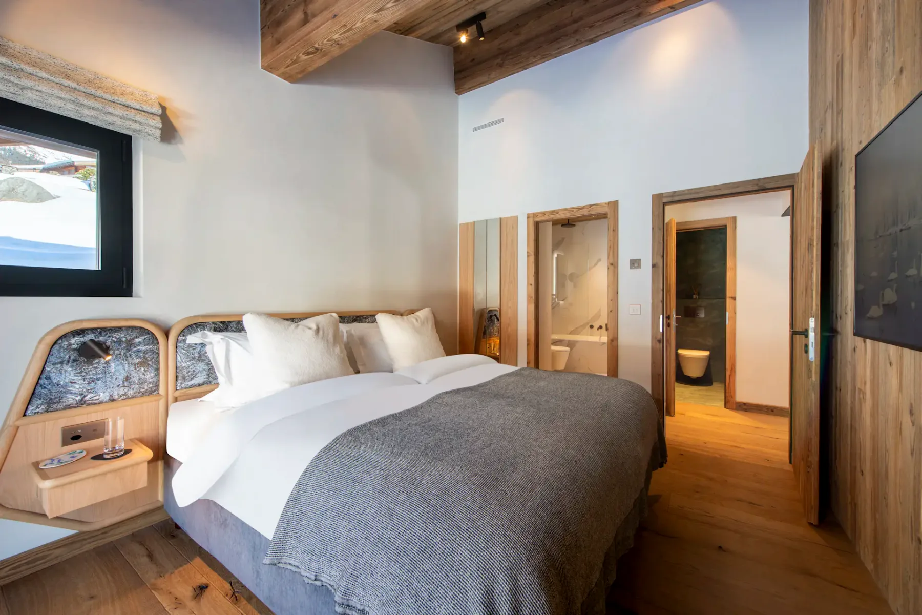Modern bedroom with wooden ceiling and floor, featuring a double bed with white linens, gray throw, and two pillows. There's a large window with curtains and a wall-mounted flat-screen TV.