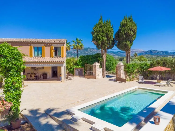 Una piscina y una terraza pavimentada junto a una casa de dos plantas, con árboles al fondo.