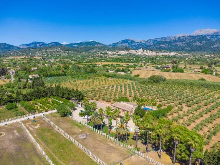 Vista aérea de una propiedad rural entre huertos, campos y montañas lejanas.