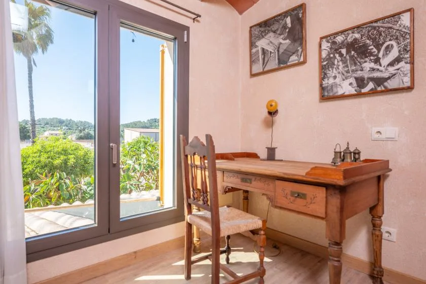 Un escritorio de madera y una silla están junto a ventanas altas con vistas a la vegetación.