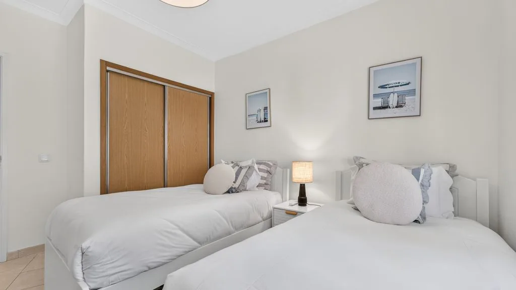 A bright bedroom with two beds, framed pictures, and a bedside lamp.