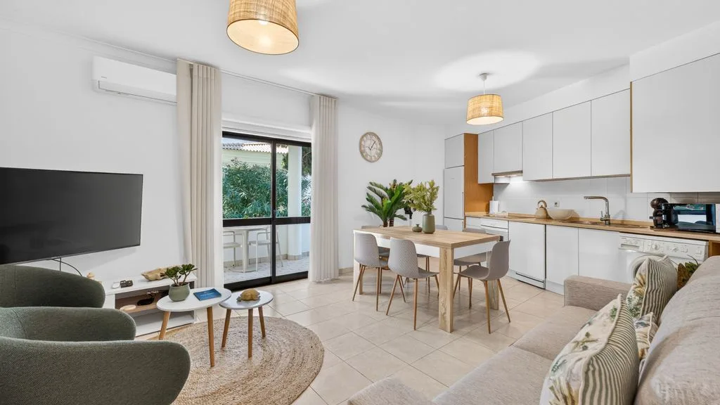 A bright living and dining area has a table beside a window and balcony doors.