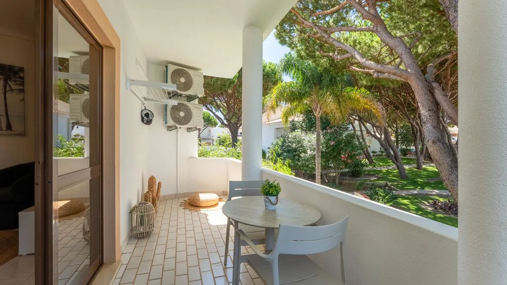 Covered balcony with a small table and chairs overlooking green trees.