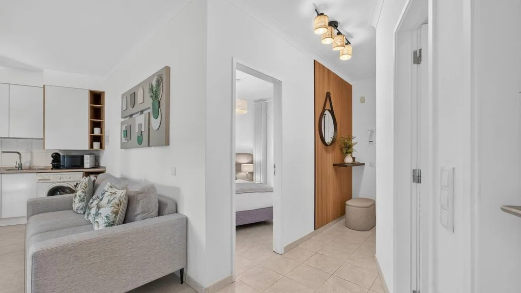 A bright apartment interior shows a seating area beside an open kitchen and hallway.