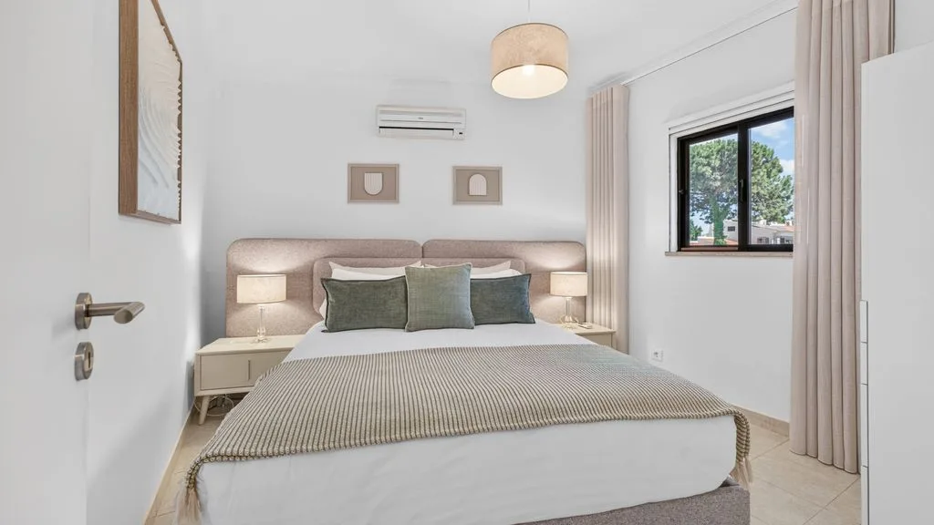 A bright bedroom with a neatly made bed and a window overlooking greenery.
