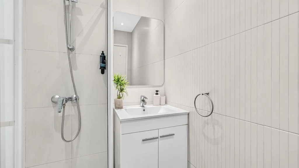 A bright bathroom features a vanity with a mirror and a wall-mounted shower.