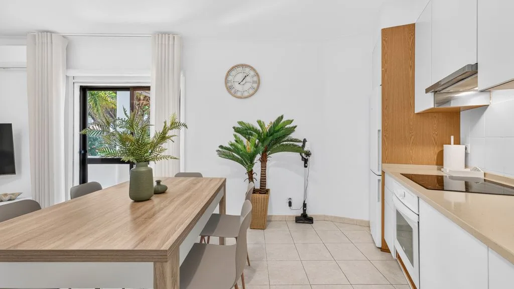 A bright dining area beside a kitchen, with a wooden table and potted plants.