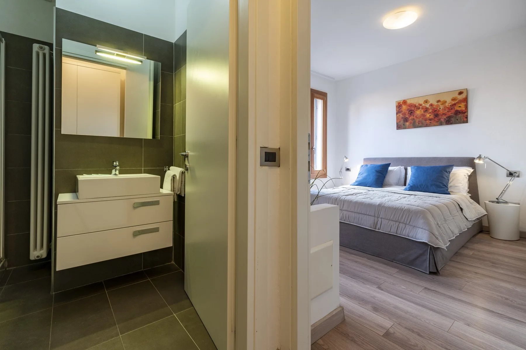 A bedroom with a bed and blue accent pillows is visible beside a bathroom vanity.