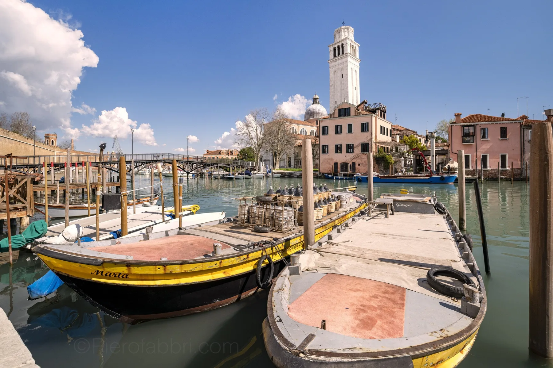 Canal-side scene with boats moored beside a wooden dock and buildings beyond.