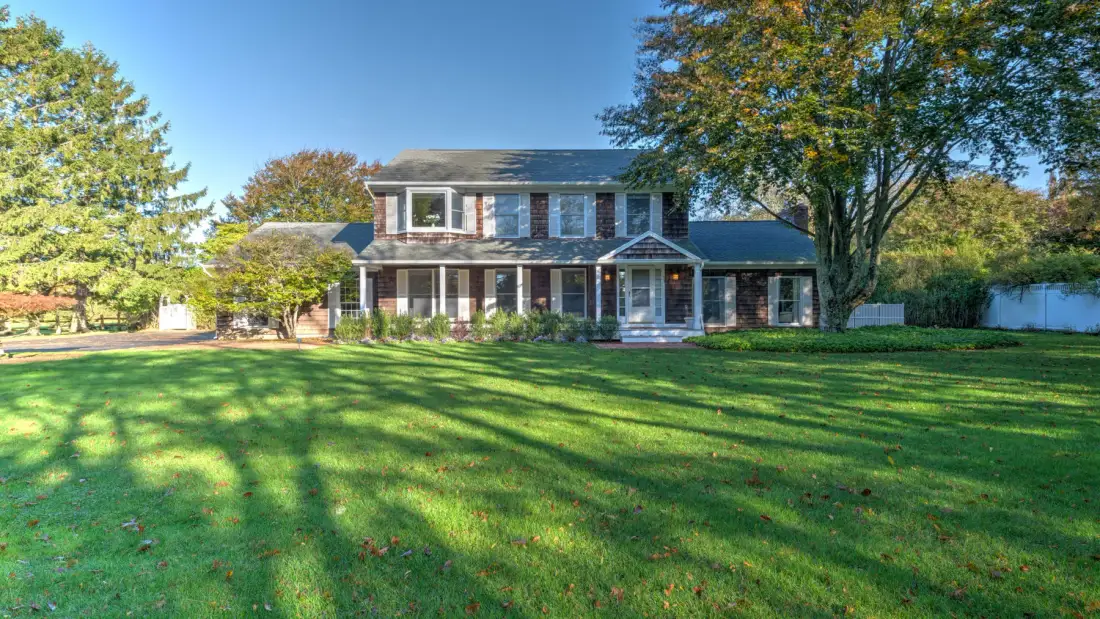 A two-story house stands behind a broad green lawn and mature trees.