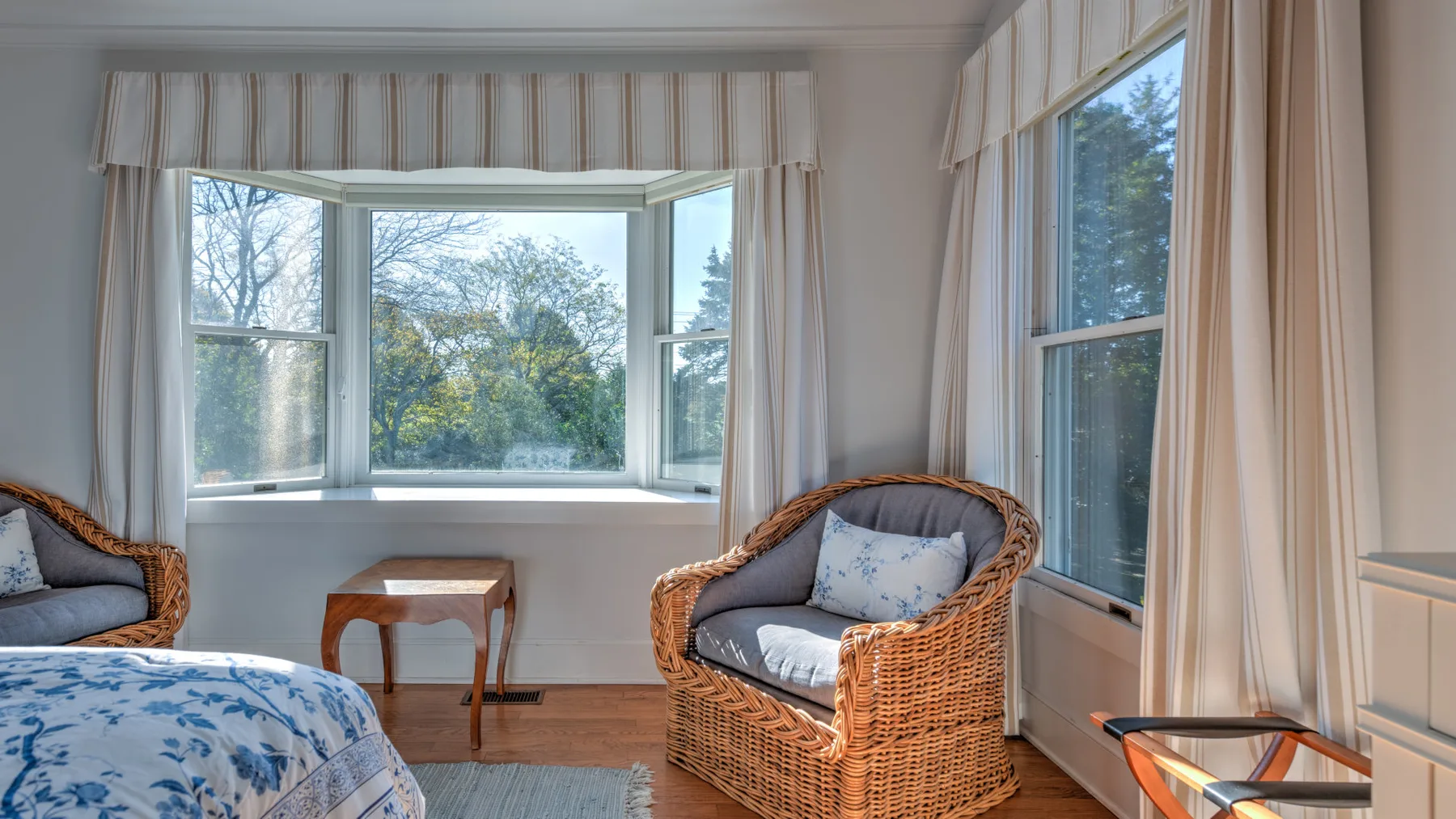 A bright bedroom with wicker seating beside large curtained windows.
