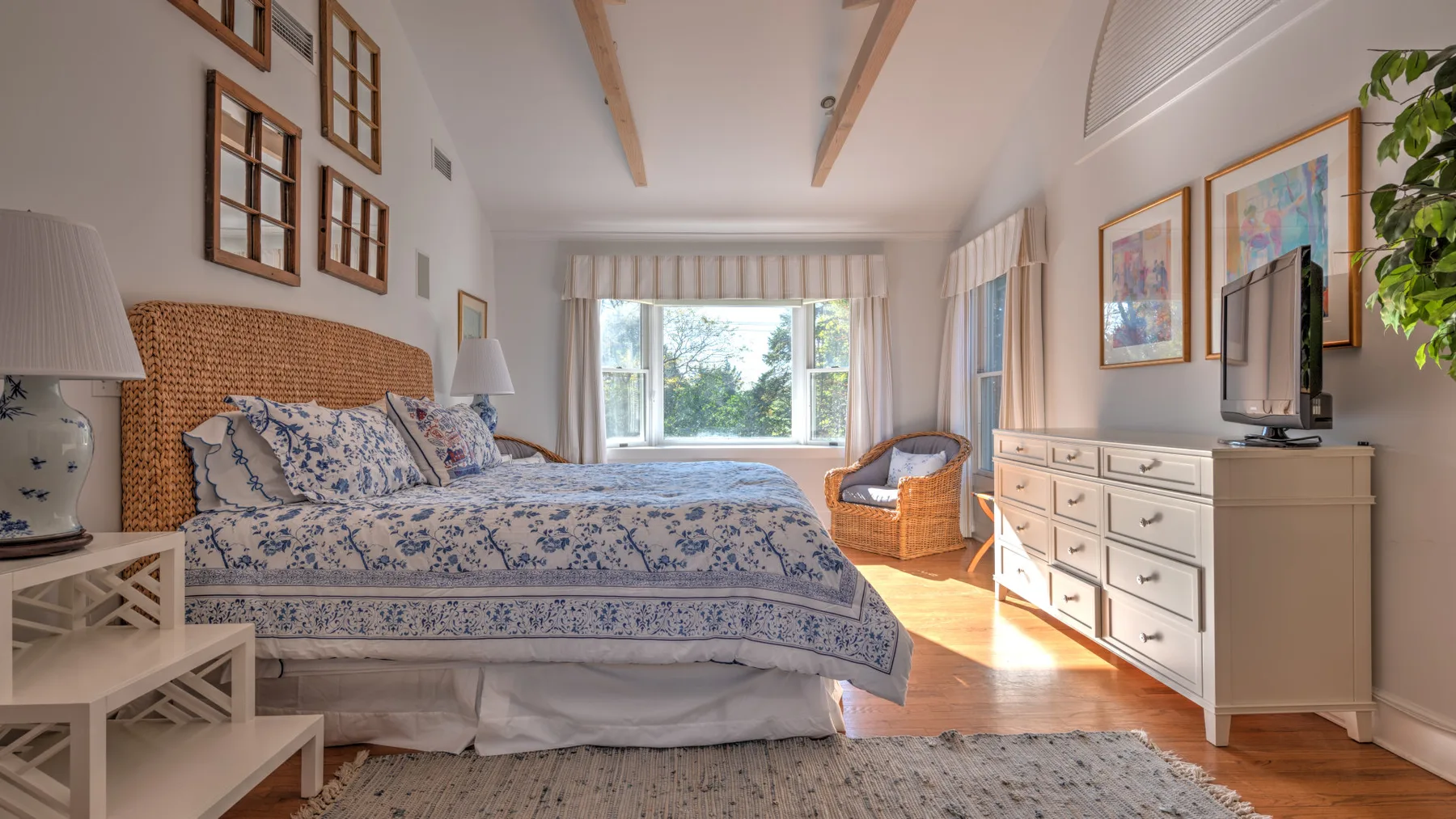 A bright bedroom with a floral bedspread, dresser, and large windows.