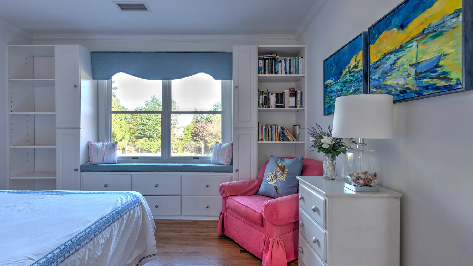 A bedroom with a bed, a pink armchair, and built-in shelving beside the window.