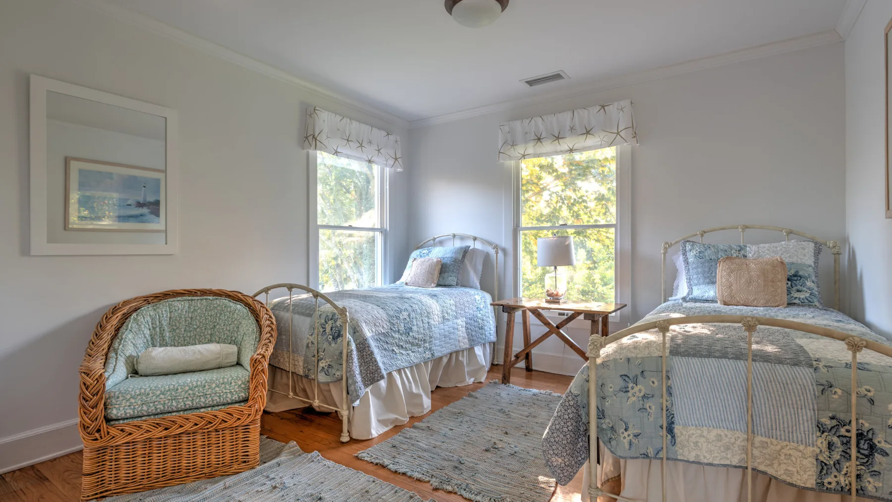 A bright bedroom with two beds, a wicker chair, and windows overlooking greenery.