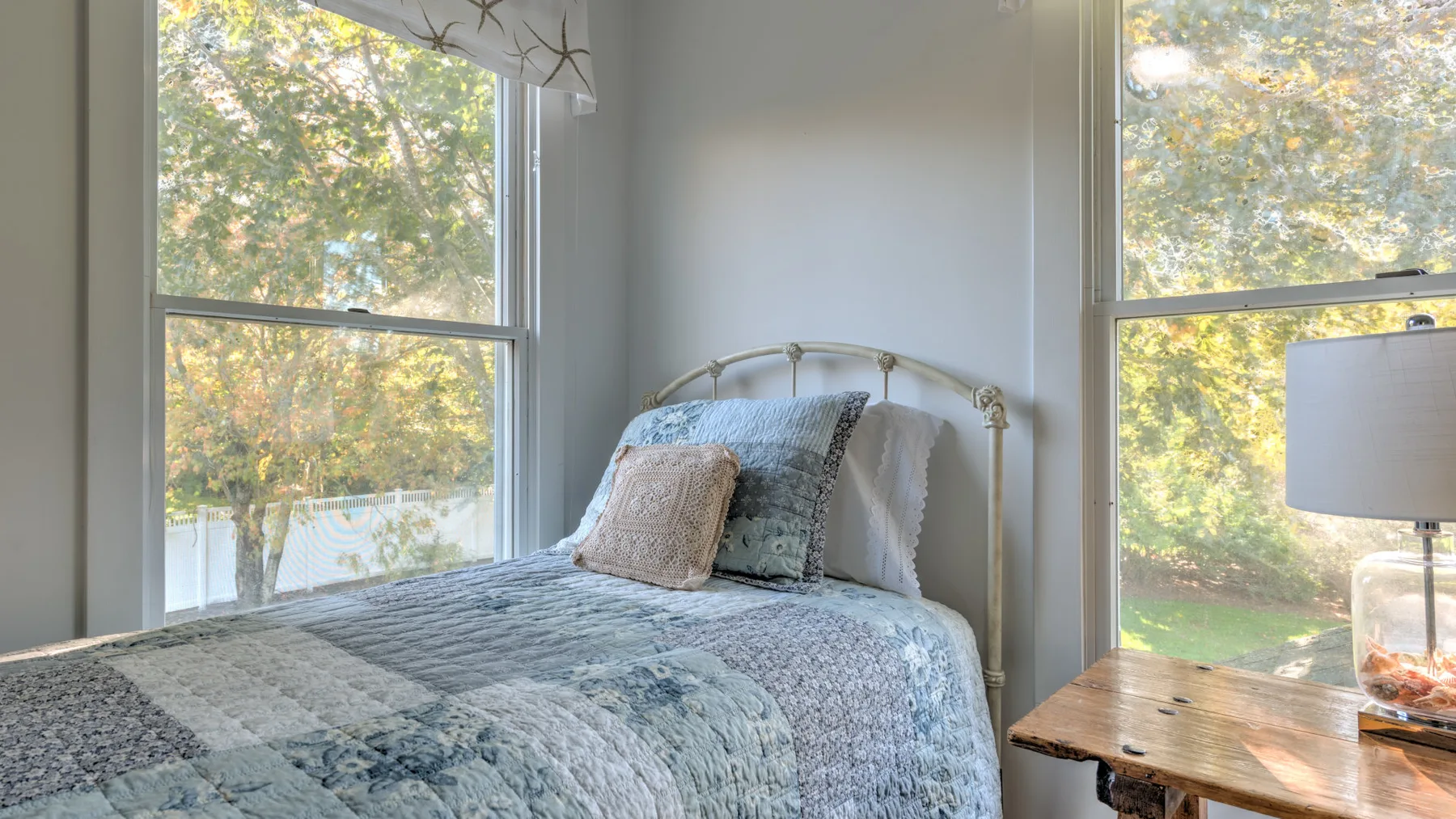 A bedroom with a bed beside large windows overlooking leafy trees.