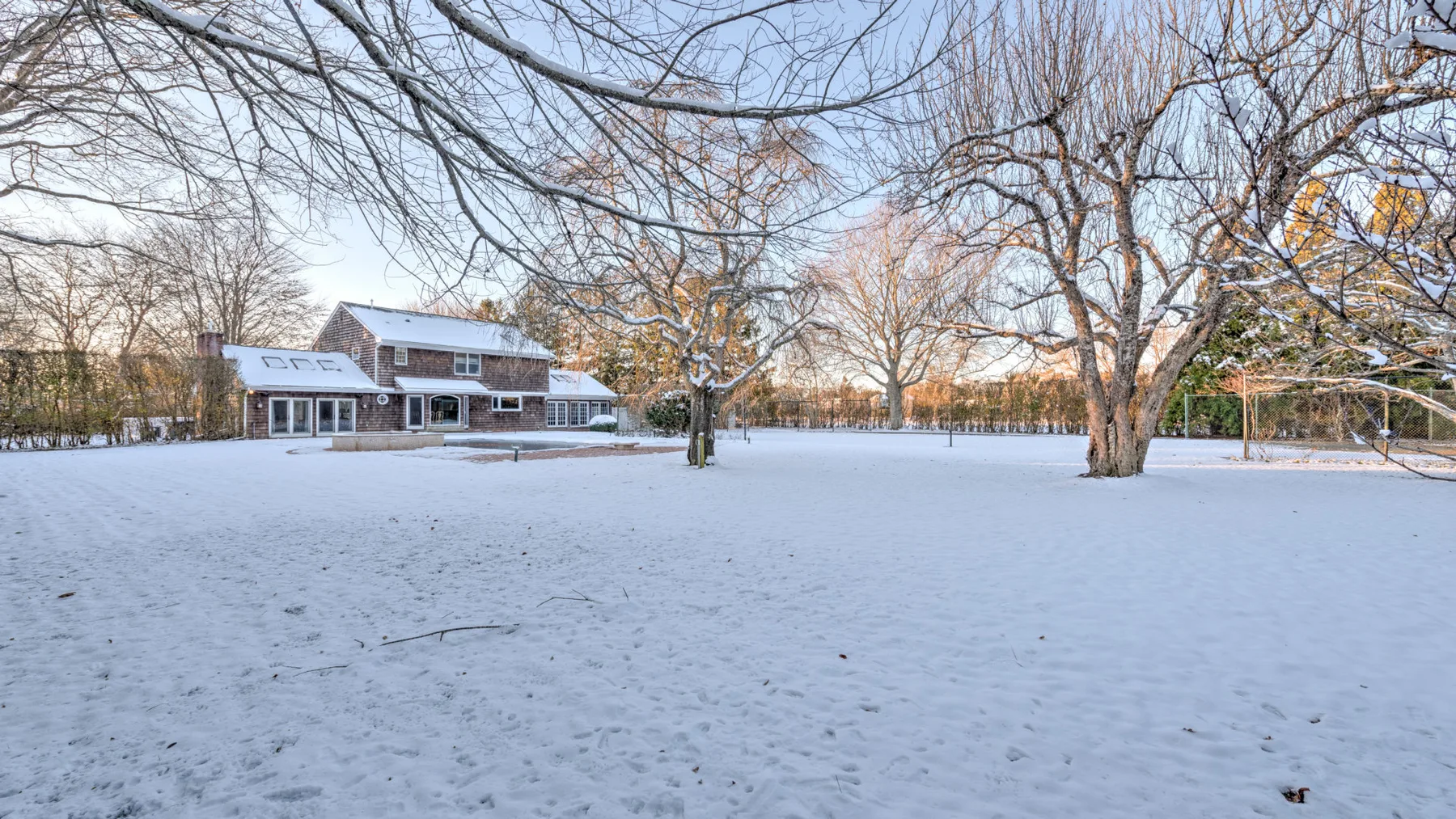 A snow-covered yard surrounds a large house and bare trees.