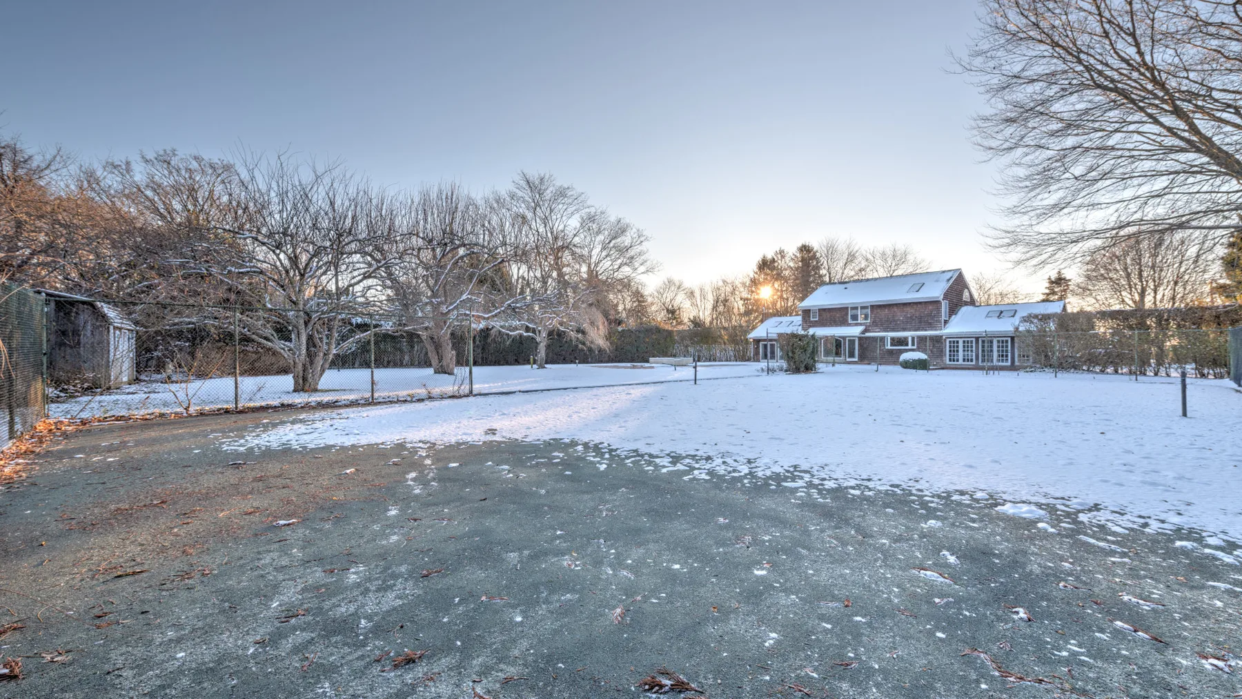 A snow-covered yard stretches toward a house and leafless trees.