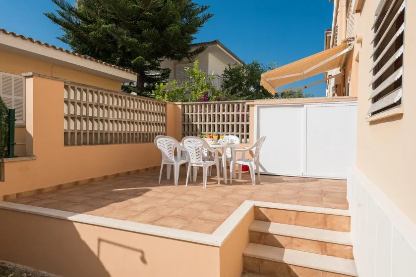 Un patio exterior de baldosas tiene asientos blancos y una mesa junto a una pantalla.
