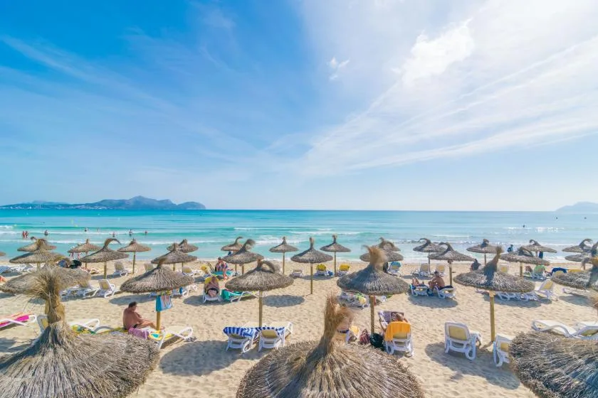 Una playa de arena con sombrillas de paja, tumbonas y agua turquesa.