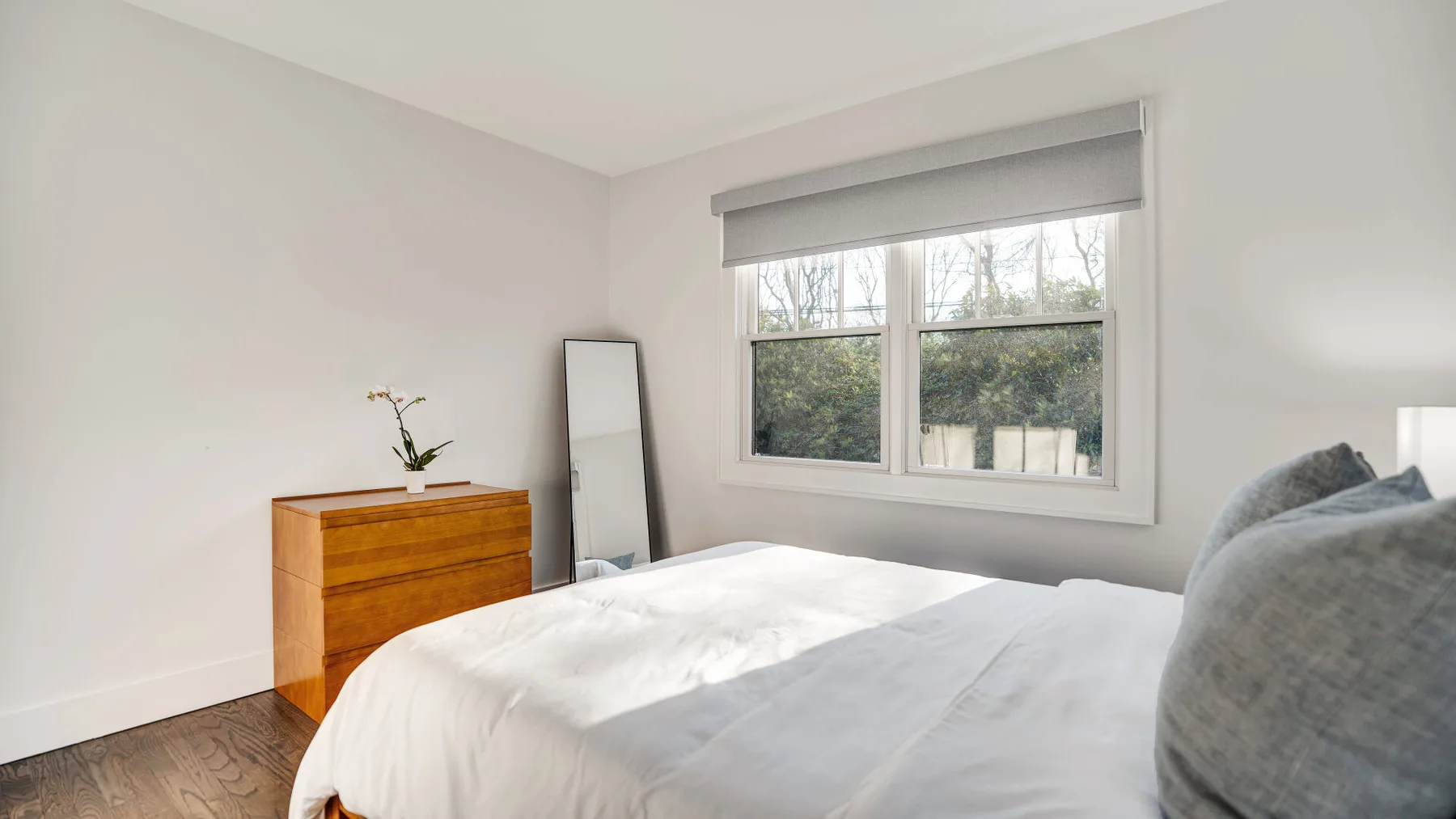 A bright bedroom with a bed, wooden dresser, and large window.