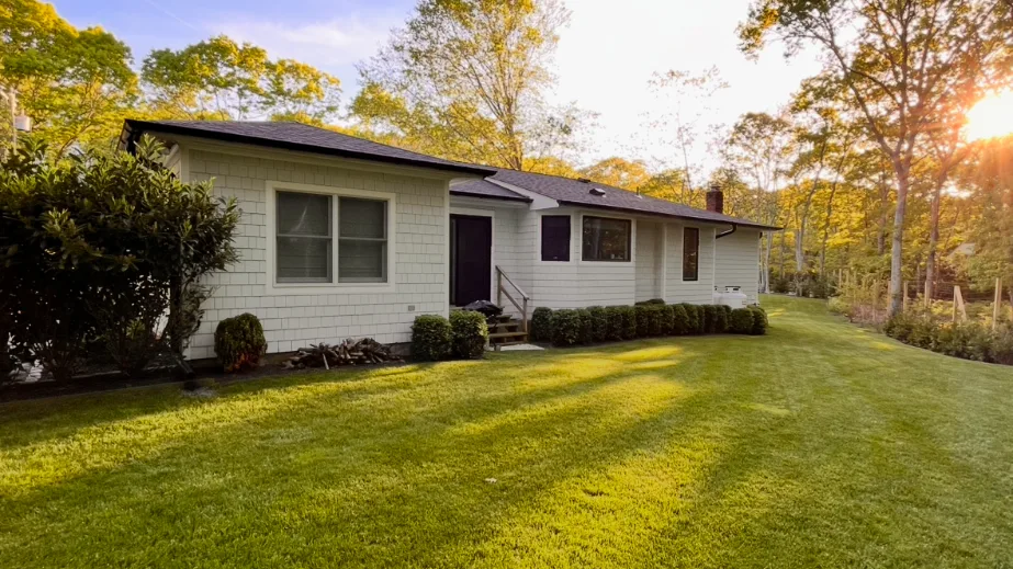 A single-story house stands beside a broad green lawn and surrounding trees.