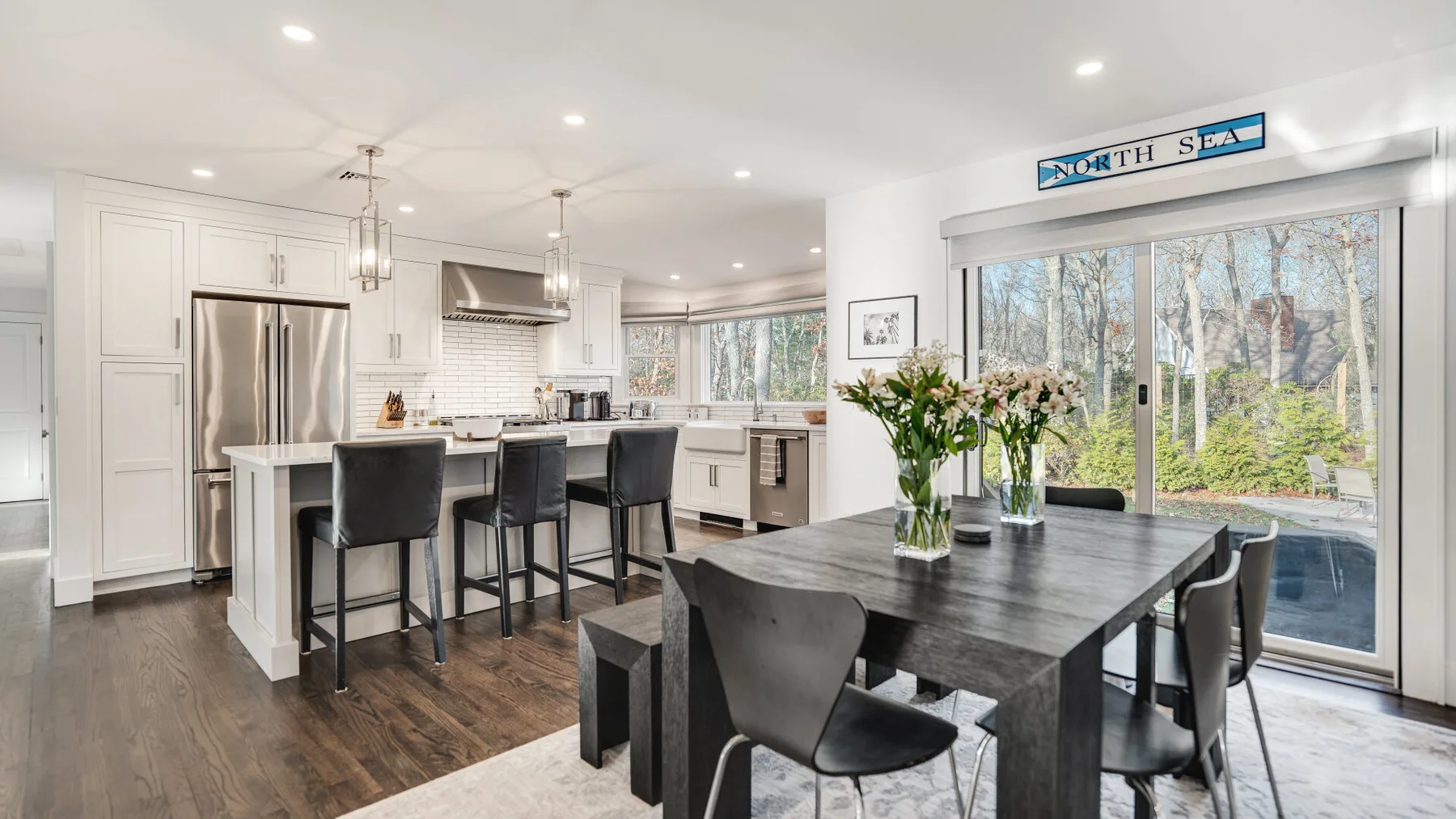 A bright open kitchen and dining area with a dark table beside large windows.