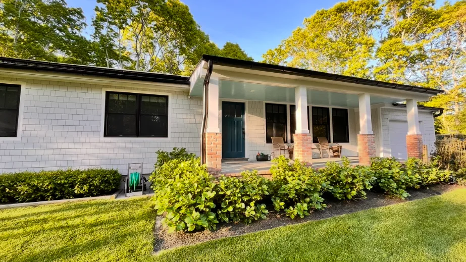 A single-story house with a covered porch stands behind shrubs and a lawn.