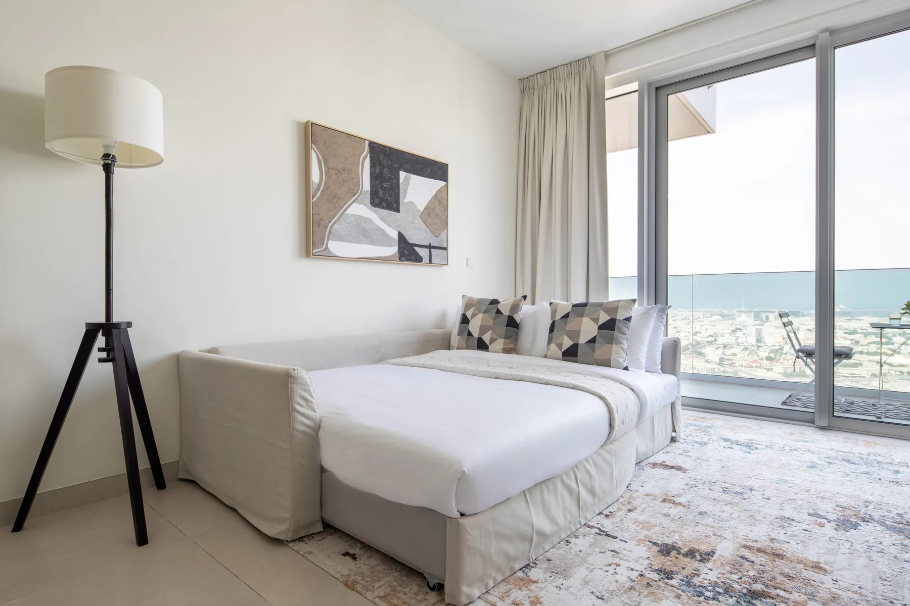 A bright bedroom with a neatly made bed beside a large window overlooking the sea.