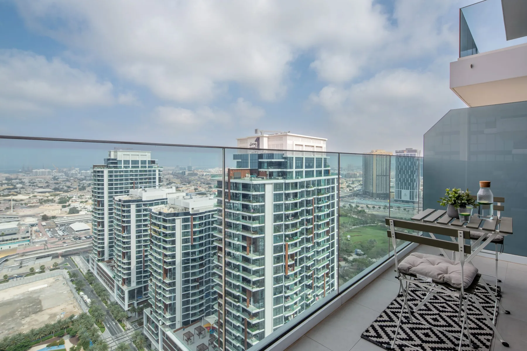 A high-rise balcony overlooks modern buildings, with a small table and seating.