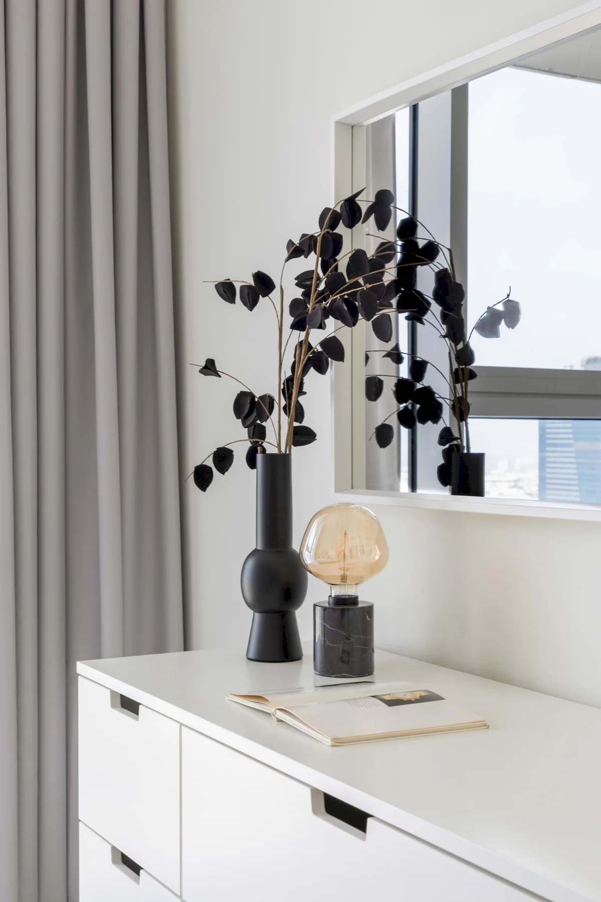 A white dresser beneath a window holds a vase, lamp, and book.