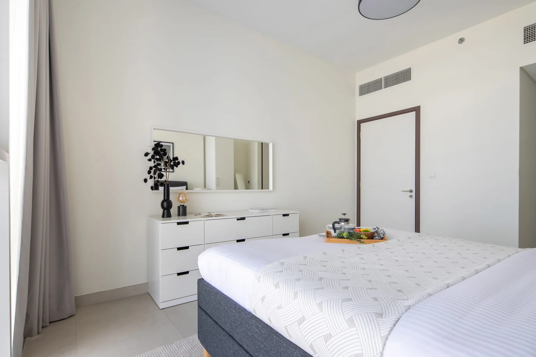 A bright bedroom with a bed, a white dresser, and a wall mirror.