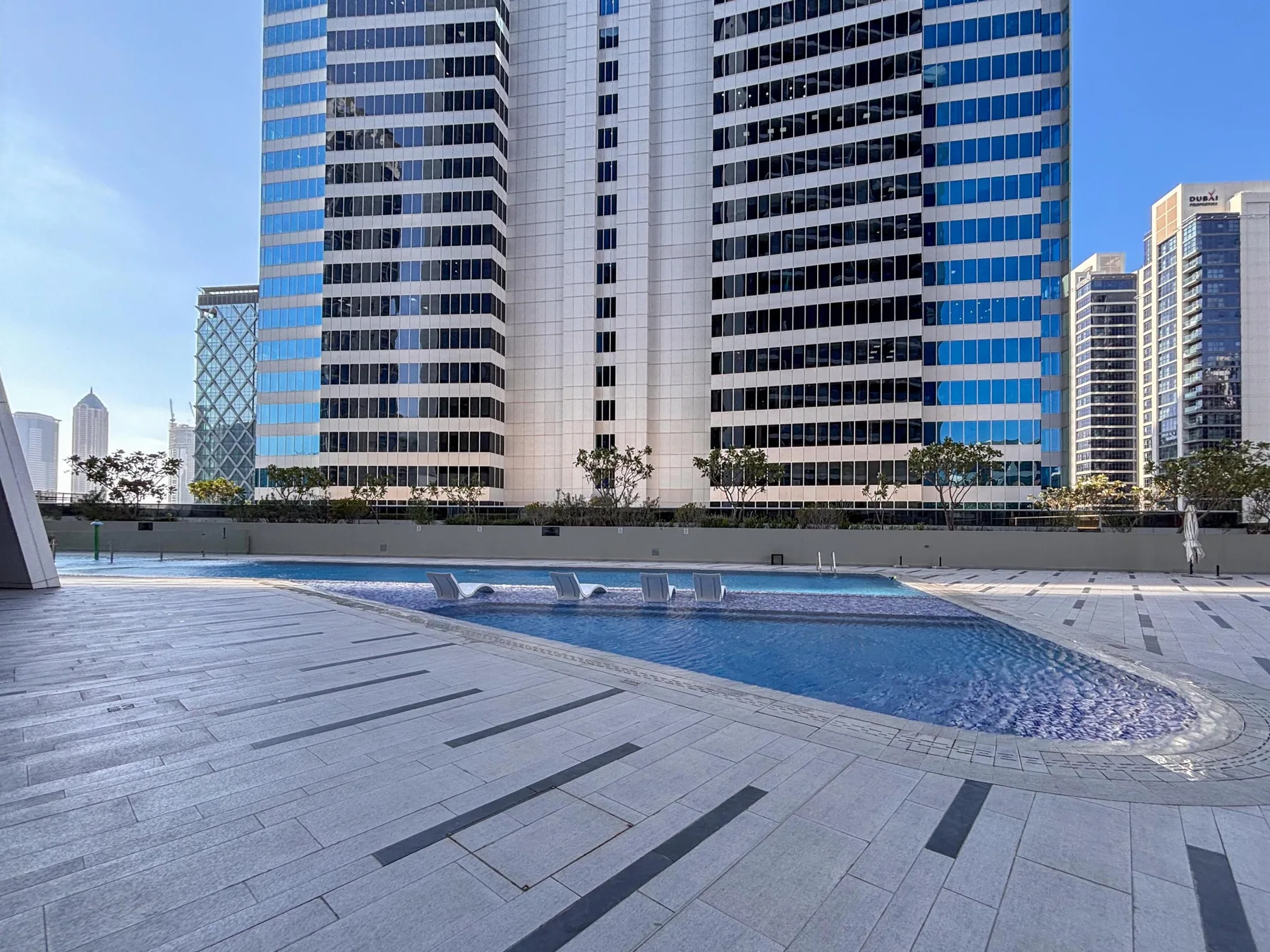 Una piscina al aire libre está junto a una terraza pavimentada, con edificios altos al fondo.