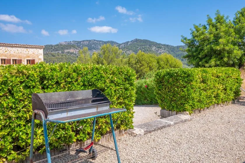 Una parrilla está junto a setos en un patio de grava, con colinas a lo lejos.