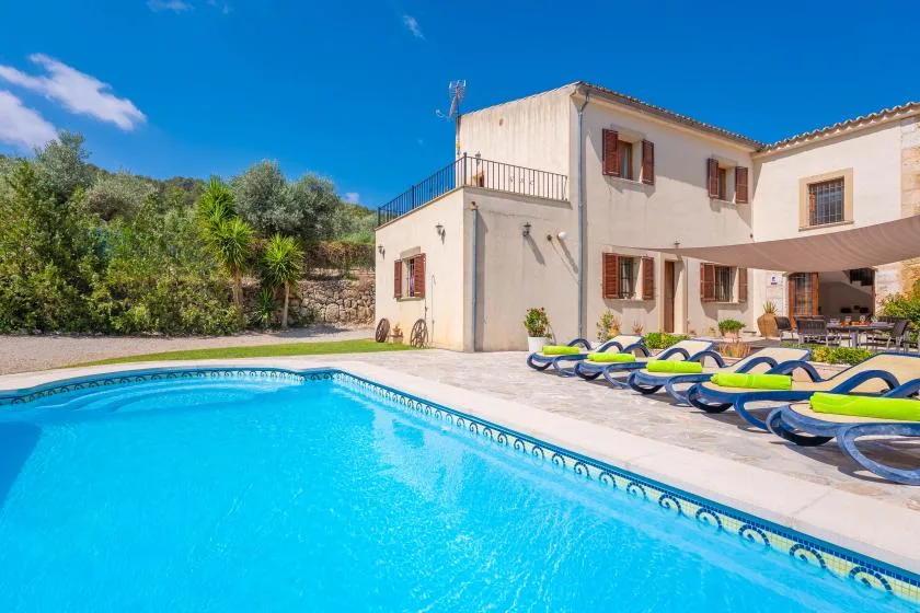 Una piscina exterior junto a una casa de color claro y filas de tumbonas.