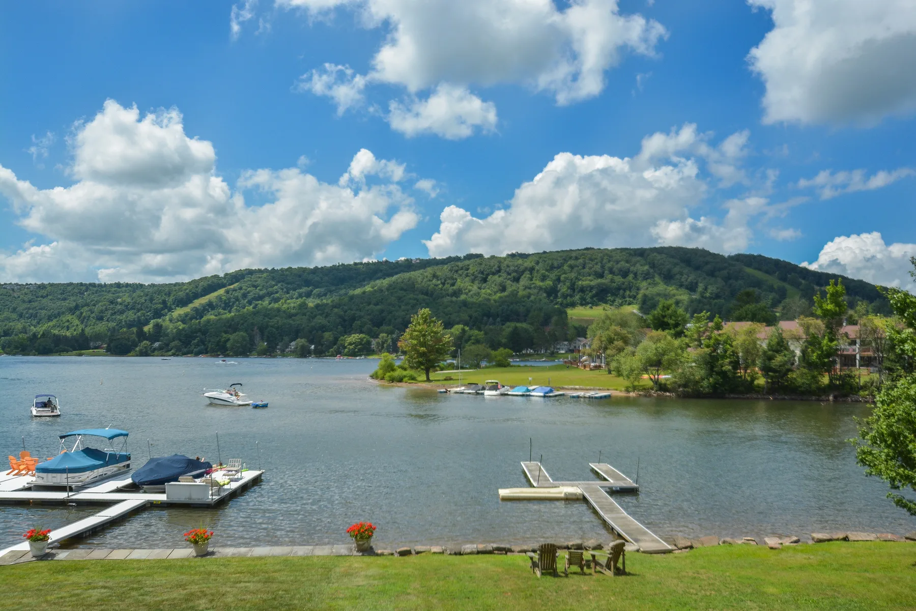A lakeside scene with boats on the water and wooded hills beneath a partly cloudy sky.