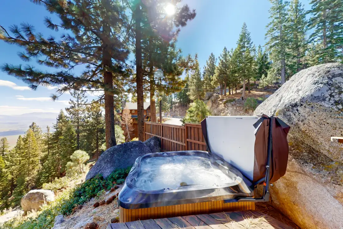 An outdoor hot tub sits on a wooden deck among pine trees, with a mountain view.