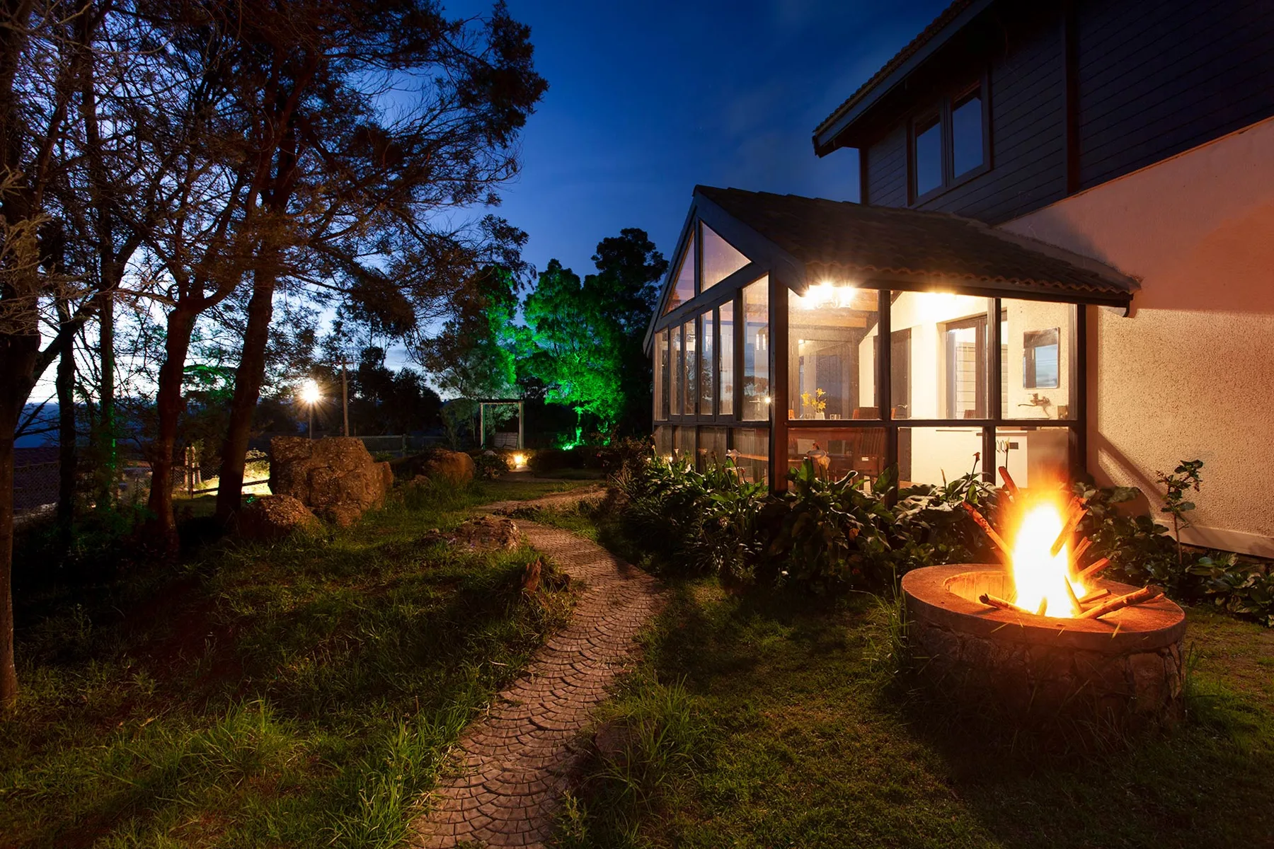 Una casa iluminada y un brasero exterior junto a un sendero de jardín al anochecer.