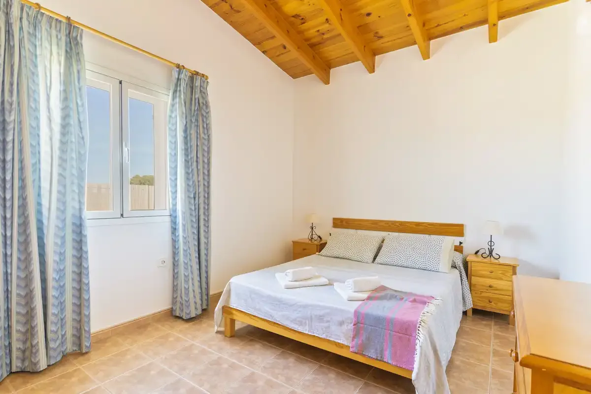 A bright bedroom with a bed, bedside tables, and a window beneath a wood ceiling.