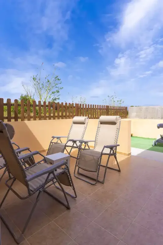 A sunny outdoor patio with reclining seating beside a low wall and wooden fence.