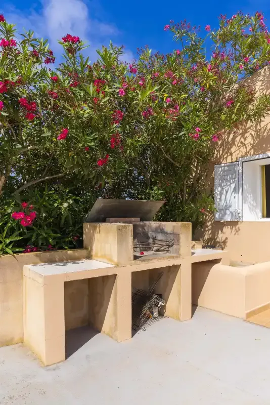 An outdoor masonry barbecue stands beneath flowering shrubs beside a window.
