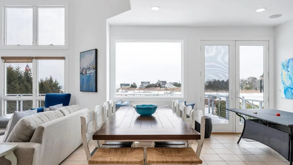 A bright dining and living area with a long table beside large windows.